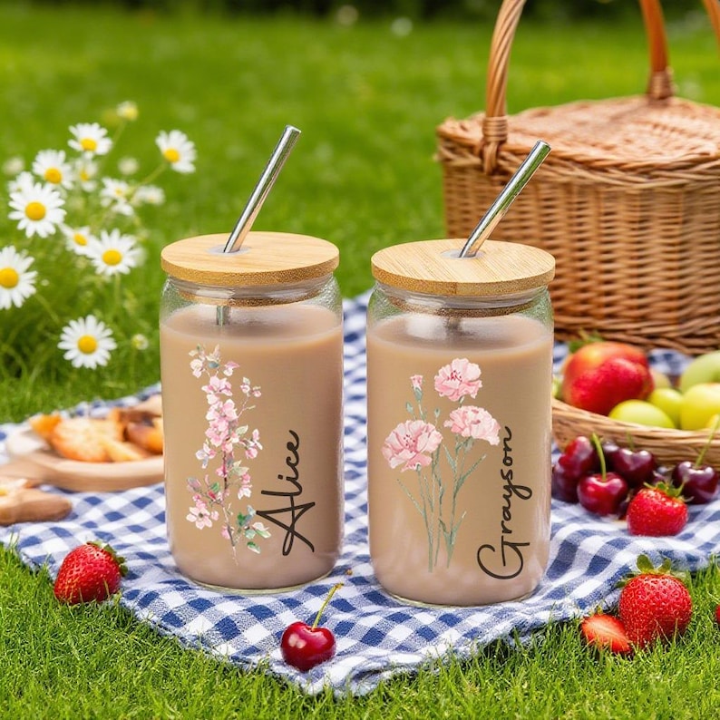 Puede incluir: Dos vasos de vidrio con tapas de madera y pajitas de metal, llenos de una bebida marr&oacute;n claro. Cada vaso presenta un dise&ntilde;o floral y un nombre: "Alice" y "Grayson". Los vasos est&aacute;n sobre una manta a cuadros azules y blancos.