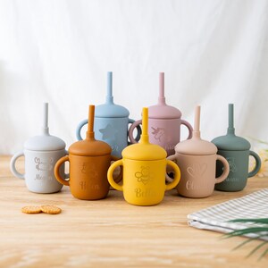 May include: A collection of colorful silicone sippy cups with lids and straws. Each cup has a different color, including blue, yellow, and pink. The cups feature engraved designs and names, such as a crab, star, bee, and unicorn. Two cookies are in the foreground.