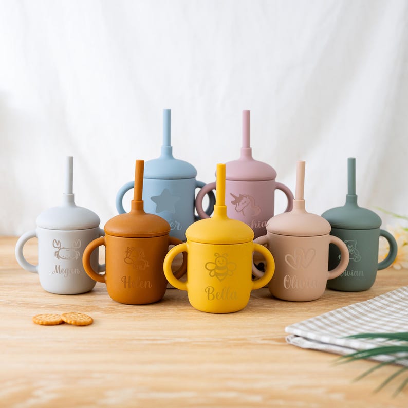 May include: A collection of colorful silicone sippy cups with lids and straws. Each cup has a different color, including blue, yellow, and pink. The cups feature engraved designs and names, such as a crab, star, bee, and unicorn. Two cookies are in the foreground.