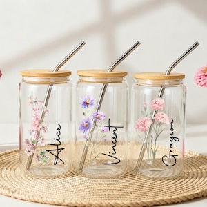 Puede incluir: Tres tarros de vidrio transparente con tapas de bamb&uacute; y pajitas de metal, cada uno decorado con dise&ntilde;os florales y nombres personalizados: Alice, Vincent y Grayson. Los tarros est&aacute;n sobre una estera tejida, con flores rosas al fondo.