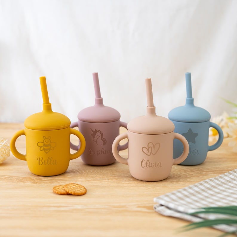 May include: Four silicone sippy cups in yellow, mauve, peach, and blue. Each cup has a lid, handles, and a straw. The yellow cup has a bee graphic and the name "Bella". The mauve cup has a unicorn graphic and the name "Sophia". The peach cup has two hearts and the name "Olivia". The blue cup has a star and the name "Carter".