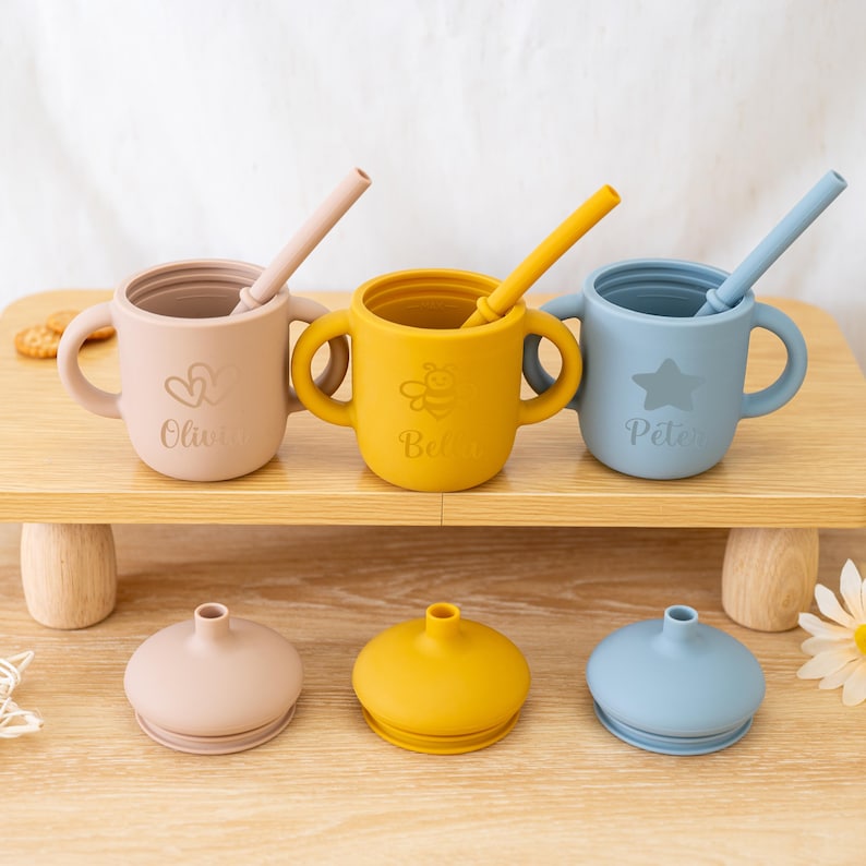 May include: Three silicone sippy cups in pastel colors: pink, yellow, and blue. Each cup has a straw and handles. The pink cup has a heart design and the name "Olivia". The yellow cup has a bee design and the name "Bela". The blue cup has a star design and the name "P&eacute;ter".