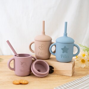 May include: Three silicone sippy cups in pink, tan, and blue, each with a lid and handles. The pink cup has a unicorn and the name "Sophia". The tan cup has hearts and the name "Olivia". The blue cup has a star and the name "Peter".