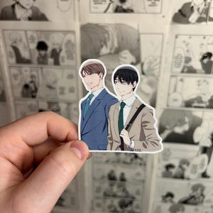 May include: A sticker featuring two anime-style male characters. One is in a blue suit with a blue tie, and the other is in a tan suit with a green tie and a black bag. The background is a wall covered in manga pages.