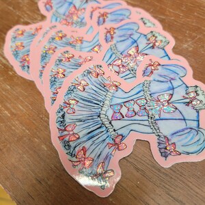 K&ouml;nnte beinhalten: Ein Stapel Aufkleber mit einem blauen Kleid-Design mit rosa Schleifen und holografischen Akzenten. Die Aufkleber haben einen rosa Rand und liegen auf einer Holzoberfl&auml;che.