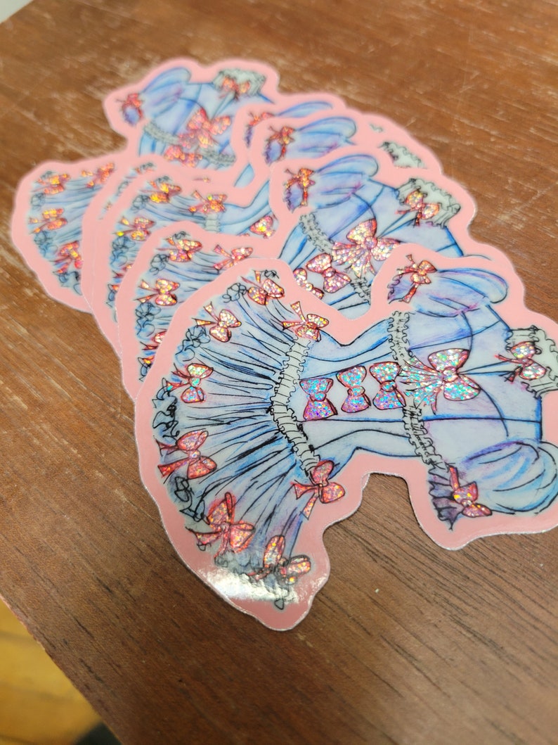 K&ouml;nnte beinhalten: Ein Stapel Aufkleber mit einem blauen Kleid-Design mit rosa Schleifen und holografischen Akzenten. Die Aufkleber haben einen rosa Rand und liegen auf einer Holzoberfl&auml;che.