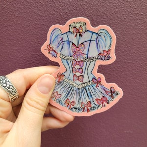 K&ouml;nnte beinhalten: Ein Aufkleber mit einem blau-wei&szlig;en Kleid-Design mit rosa Schleifen und Akzenten. Der Aufkleber hat einen rosa Rand und wird von einer Hand vor einem lila Hintergrund gehalten. Das Kleid hat einen R&uuml;schenrock und Puff&auml;rmel.