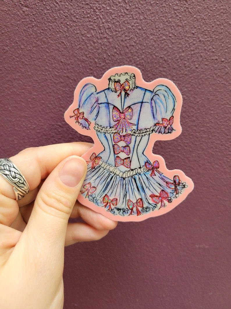K&ouml;nnte beinhalten: Ein Aufkleber mit einem blau-wei&szlig;en Kleid-Design mit rosa Schleifen und Akzenten. Der Aufkleber hat einen rosa Rand und wird von einer Hand vor einem lila Hintergrund gehalten. Das Kleid hat einen R&uuml;schenrock und Puff&auml;rmel.