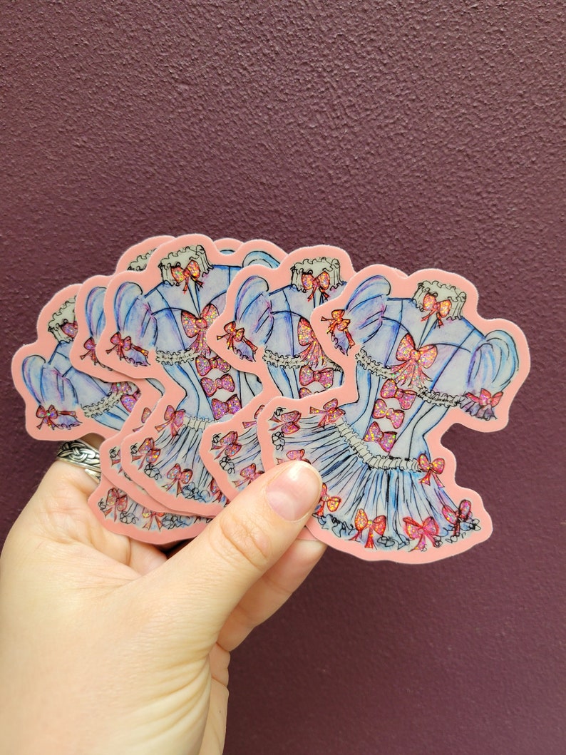 K&ouml;nnte beinhalten: Eine Ansammlung von Aufklebern mit einem hellblauen Kleid-Design mit rosa Schleifen und Akzenten. Die Aufkleber haben einen rosa Rand und sind vor einem dunkelvioletten Hintergrund zu sehen.