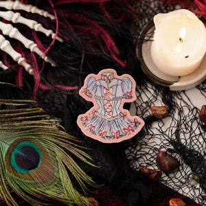 K&ouml;nnte beinhalten: Ein Aufkleber mit einem blau-rosa Korsett-Design mit Schleifen, vor einem Hintergrund aus schwarzem Netz, Federn und einer Pfauenfeder. Eine brennende Kerze und eine wei&szlig;e Skeletthand sind ebenfalls zu sehen.