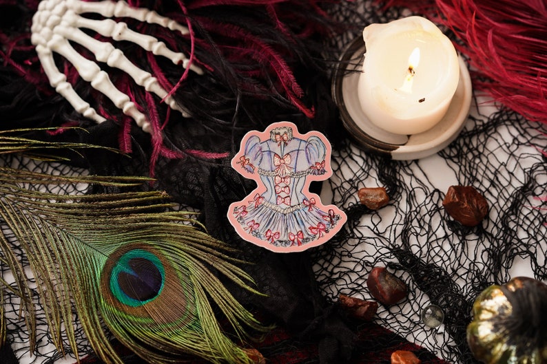 K&ouml;nnte beinhalten: Ein Aufkleber mit einem blau-rosa Korsett-Design mit Schleifen, vor einem Hintergrund aus schwarzem Netz, Federn und einer Pfauenfeder. Eine brennende Kerze und eine wei&szlig;e Skeletthand sind ebenfalls zu sehen.