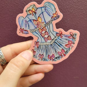 K&ouml;nnte beinhalten: Ein Aufkleber mit einem hellblauen Kleid-Design mit rosa Schleifen und Glitzerakzenten. Der Aufkleber hat einen rosa Rand und wird vor einem lila Hintergrund gehalten. Das Kleid hat einen ger&uuml;schten Rock und Puff&auml;rmel.