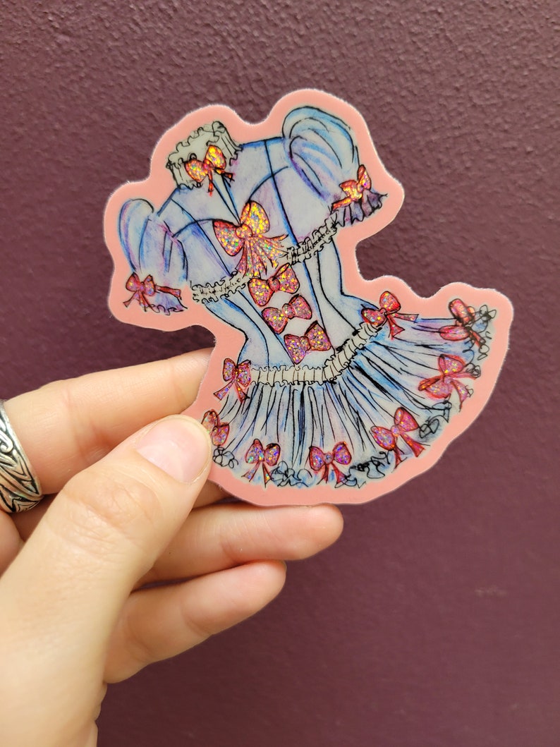 K&ouml;nnte beinhalten: Ein Aufkleber mit einem hellblauen Kleid-Design mit rosa Schleifen und Glitzerakzenten. Der Aufkleber hat einen rosa Rand und wird vor einem lila Hintergrund gehalten. Das Kleid hat einen ger&uuml;schten Rock und Puff&auml;rmel.