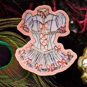 K&ouml;nnte beinhalten: Ein Aufkleber mit einer Aquarellillustration eines hellblauen Kleides mit rosa Schleifen. Das Kleid hat ein korsettartiges Oberteil und einen ger&uuml;schten Rock. Rote Schleifen zieren die &Auml;rmel, das Oberteil und den Rock. Der Aufkleber ist rosa umrandet.