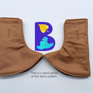 May include: A pair of brown fabric boot covers featuring a vertical buttonhole on the upper section and a flared base. Between the two covers is a purple graphic containing a yellow leaf and two light blue birds, with the text: This is a stock photo of this fabric pattern.