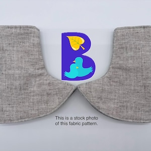 May include: Two grey linen-textured fabric pieces shaped like boot covers or cuffs, positioned symmetrically. Between them is a purple letter B graphic featuring a yellow leaf and two light blue birds. Text below reads: This is a stock photo of this fabric pattern.