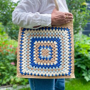 Könnte beinhalten: Eine quadratische Tragetasche mit gehäkelter Frontplatte. Das Häkeldesign zeigt ein zentrales blaues Quadrat, das von konzentrischen Ringen aus Beige, Hellblau und Weiß umgeben ist. Die Tasche hat einen Körper aus naturfarbenem Jute und einen passenden Schultergurt.