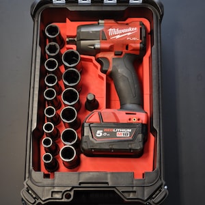 Puede incluir: Un juego de llaves de impacto y vasos Milwaukee FUEL rojo y negro en una caja negra. El juego incluye una variedad de vasos y una batería M18 REDLITHIUM de 5,0 Ah. La caja está abierta, revelando las herramientas dispuestas en un interior rojo.
