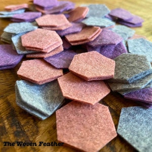 May include: A pile of felt hexagon shapes in shades of purple, pink, and gray. The hexagons are stacked on top of each other and are arranged in a random pattern. The text "The Woven Feather" is visible in the bottom left corner of the image.