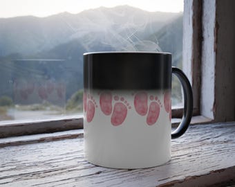 May include: A black and white color-changing mug with a row of pink baby footprints. The mug is filled with a hot beverage, creating steam. The mug sits on a weathered, white wooden windowsill.