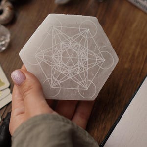 Può includere: Un cristallo di selenite esagonale, bianco sporco, con un disegno geometrico inciso sulla superficie. Il cristallo è tenuto in mano, con altri cristalli e oggetti sullo sfondo. Il disegno presenta linee e cerchi che si intersecano.