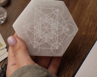 Puede incluir: Un cristal de selenita hexagonal, blanquecino, con un diseño geométrico grabado en su superficie. El cristal se sostiene en una mano, con otros cristales y objetos en el fondo. El diseño presenta líneas y círculos que se cruzan.
