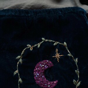 May include: Dark teal velvet fabric with an embroidered crescent moon and star design. The moon is a deep purple, embellished with beads. A gold-colored star and a leafy vine encircle the moon. The background is a dark teal color.