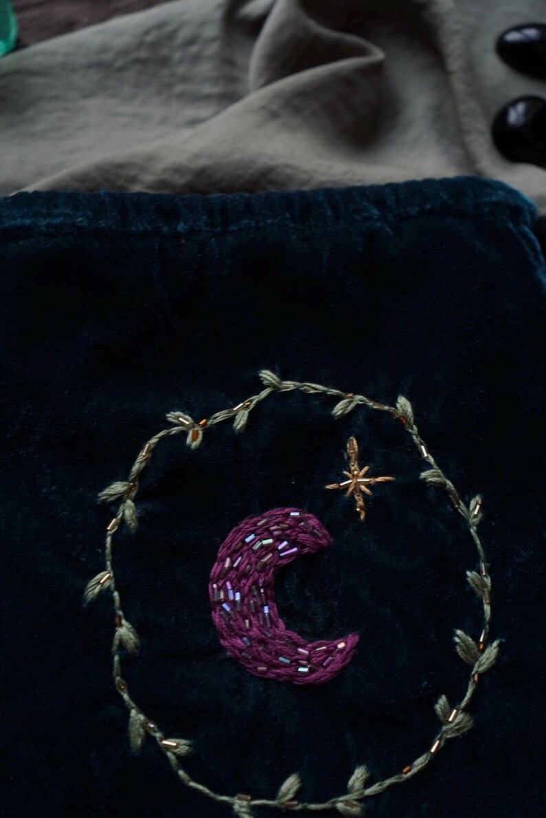 May include: Dark teal velvet fabric with an embroidered crescent moon and star design. The moon is a deep purple, embellished with beads. A gold-colored star and a leafy vine encircle the moon. The background is a dark teal color.