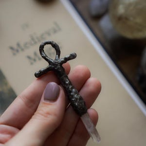 Può includere: Un ciondolo ankh in metallo scuro e testurizzato con finitura martellata e una punta di cristallo trasparente alla base. L'oggetto presenta un occhiello sulla parte superiore e bracci orizzontali, simile all'antico simbolo della croce egizia.