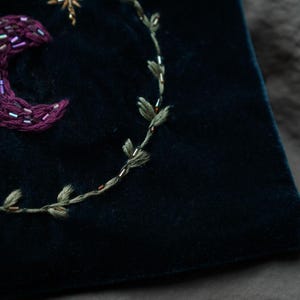 May include: Close-up of a dark blue velvet fabric with embroidered celestial designs. Features a crescent moon, star, and a leafy vine, all in shades of purple, gold, and green. The embroidery is detailed and textured.