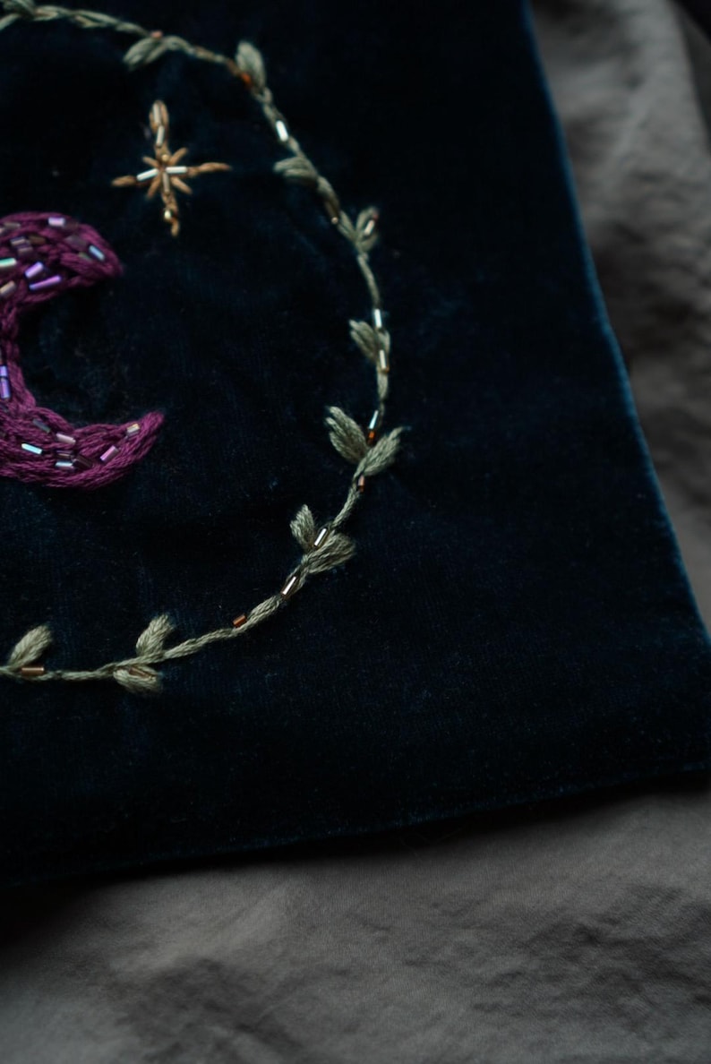 May include: Close-up of a dark blue velvet fabric with embroidered celestial designs. Features a crescent moon, star, and a leafy vine, all in shades of purple, gold, and green. The embroidery is detailed and textured.