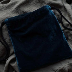 May include: A dark teal velvet drawstring pouch. The pouch is rectangular with rounded corners and a black drawstring closure. The velvet has a soft, plush texture. The background is a muted olive green fabric.