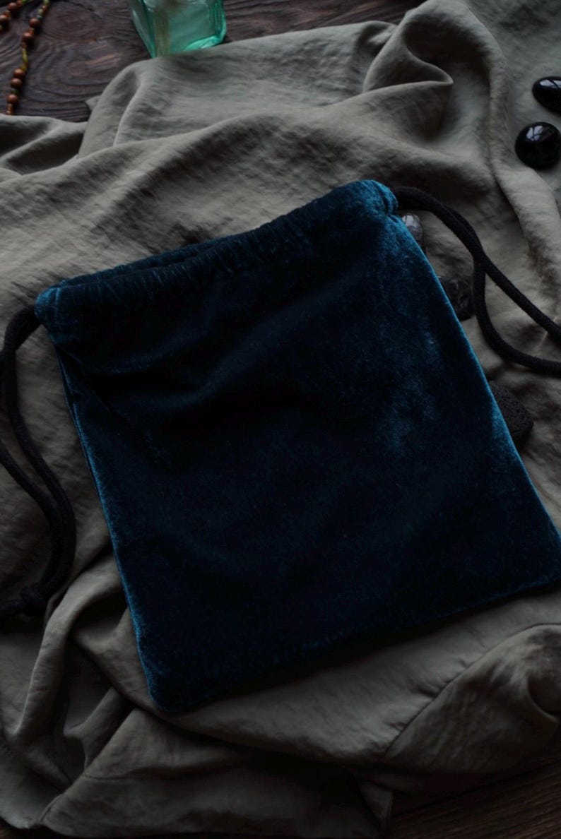 May include: A dark teal velvet drawstring pouch. The pouch is rectangular with rounded corners and a black drawstring closure. The velvet has a soft, plush texture. The background is a muted olive green fabric.