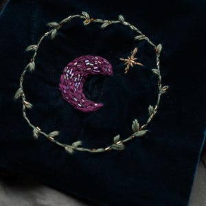 May include: Dark teal velvet pouch with embroidered celestial design. Features a crescent moon in shades of purple, a gold star, and a green leafy vine. The pouch is a small, soft bag, perfect for storing small items.