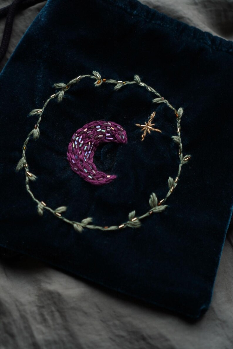 May include: Dark teal velvet pouch with embroidered celestial design. Features a crescent moon in shades of purple, a gold star, and a green leafy vine. The pouch is a small, soft bag, perfect for storing small items.