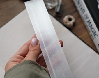 May include: A rectangular, translucent selenite crystal wand is held in a hand. The wand is smooth and appears to be about 8 inches long. The background includes a book, small vials, and other crystals, suggesting a spiritual or metaphysical theme.