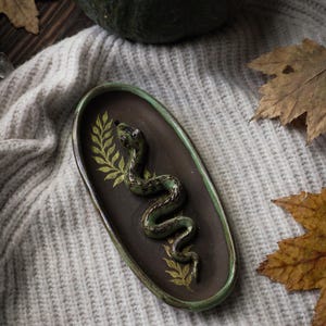 Puede incluir: Plato ovalado de cerámica con un diseño de serpiente verde en relieve. El plato es de color marrón oscuro con un borde verde y detalles de hojas doradas. Una calabaza verde oscuro y hojas de otoño en el fondo.