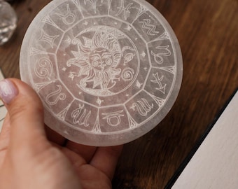May include: A circular, translucent crystal coaster featuring etched zodiac symbols and a sun design. The coaster is held in a hand, with a wooden surface in the background. The coaster is a light, neutral colour.