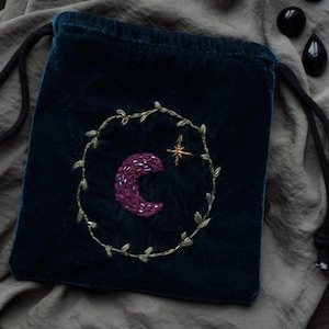 May include: A dark teal velvet drawstring bag with an embroidered design. The design features a purple crescent moon and a gold star within a gold leaf wreath. The bag is on a gray fabric surface.