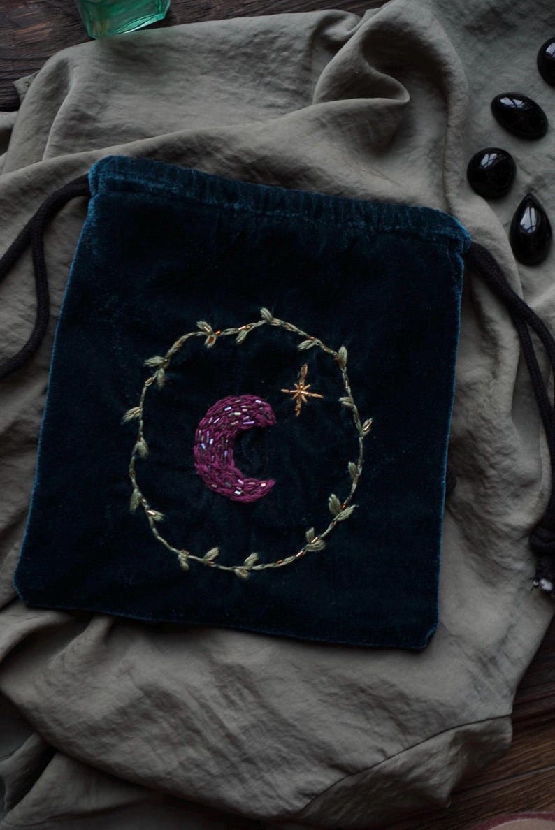 May include: A dark teal velvet drawstring bag with an embroidered design. The design features a purple crescent moon and a gold star within a gold leaf wreath. The bag is on a gray fabric surface.