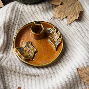 May include: A ceramic candle holder in shades of brown and green. The holder has a circular base with a central candle cup and decorative leaf and insect designs. The overall aesthetic suggests a rustic, autumnal style.