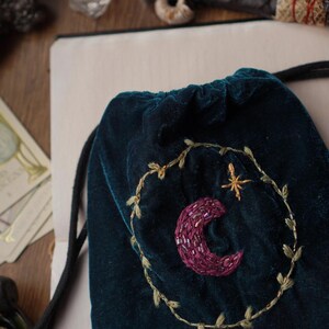 May include: A dark teal velvet drawstring bag with an embroidered crescent moon and star design. The moon is a deep purple, and the star is gold. The bag is decorated with a gold embroidered wreath.