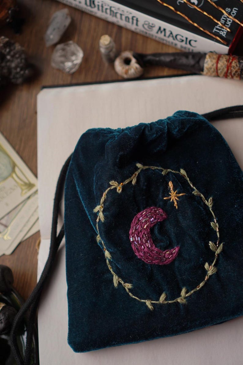 May include: A dark teal velvet drawstring bag with an embroidered crescent moon and star design. The moon is a deep purple, and the star is gold. The bag is decorated with a gold embroidered wreath.