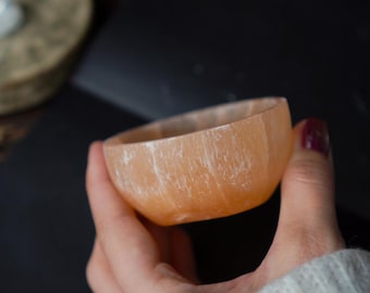May include: A small, orange-hued bowl with a textured surface. The bowl is held in a hand, and the background is dark. The bowl appears to be made of a natural material, possibly stone or crystal.