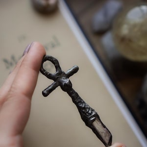 May include: A dark metallic ankh pendant featuring a textured, uneven surface and a clear, pointed crystal shard embedded at the base. The cross-shaped charm has a circular loop at the top and a rugged, hand-crafted appearance throughout.