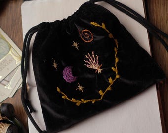May include: Black velvet drawstring bag with embroidered celestial designs, including a crescent moon, stars, and a sun. The bag is open, revealing a blank page. The background includes a book with the title "Cult, Witchcraft & Magic".