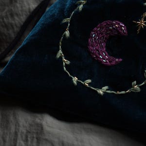 May include: A dark teal velvet drawstring bag with embroidered celestial details. A crescent moon, a star, and a leafy vine are embroidered on the bag. The bag is on a gray surface.