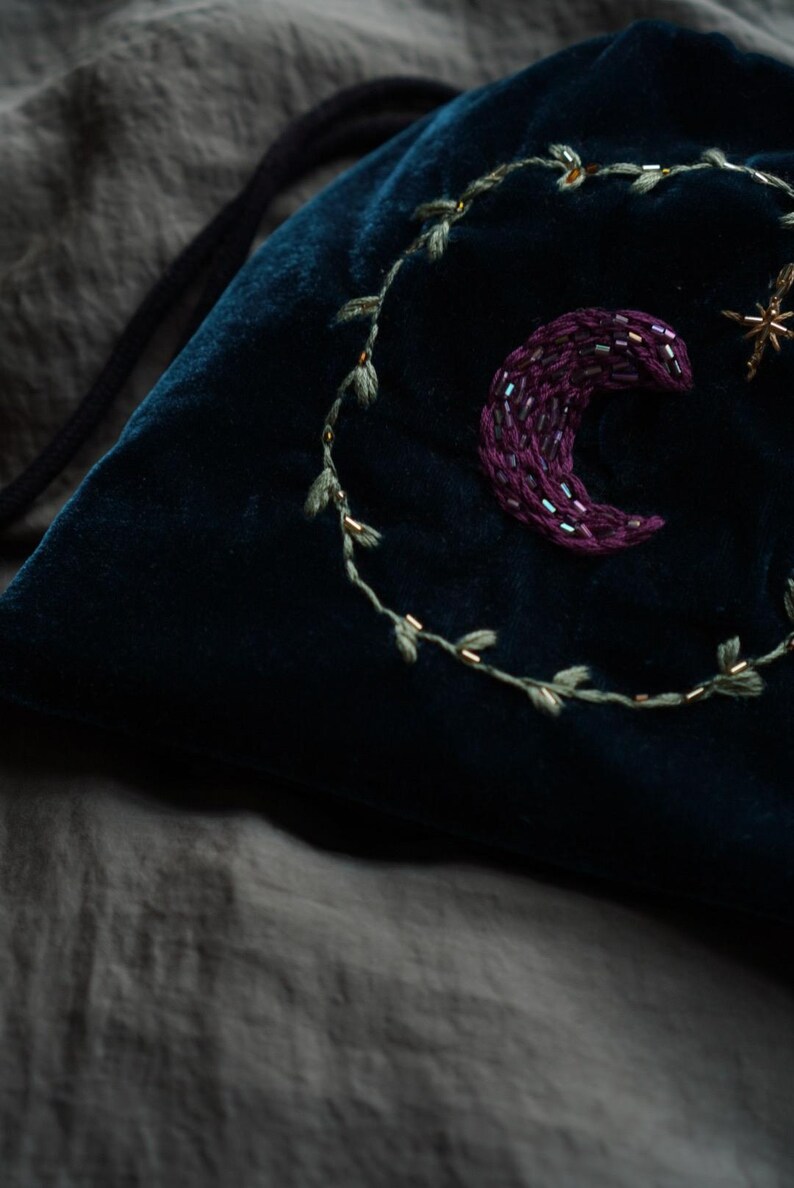 May include: A dark teal velvet drawstring bag with embroidered celestial details. A crescent moon, a star, and a leafy vine are embroidered on the bag. The bag is on a gray surface.