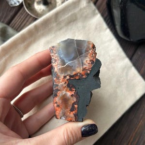 Könnte beinhalten: Eine Hand hält einen rohen, natürlichen Achatstein. Der Stein weist eine Mischung aus Farben auf, darunter Grau, Orange und Schwarz. Der Achat hat eine raue, strukturierte Oberfläche und eine einzigartige, organische Form. Der Hintergrund umfasst ein neutrales Tuch und andere Objekte.