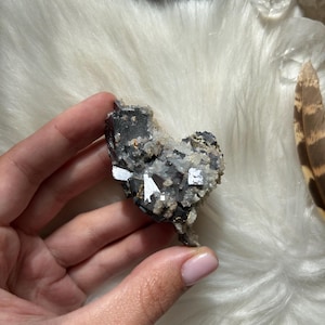 Può includere: Un gruppo a forma di cuore di cristalli metallici grigio scuro e argento con piccole formazioni cristalline di colore chiaro. Il campione minerale è tenuto in mano su uno sfondo di tessuto bianco e soffice, con una piuma e altri cristalli visibili.